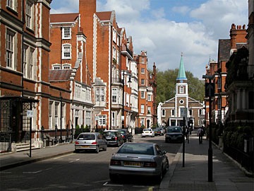 streetscape in London with brick buildings trimmed in stone on either side of the street and a church with a weathered copper steeple at the end