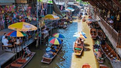 People sit on both sides of a river in Thailand. Wooden boats transport people and goods on the water.