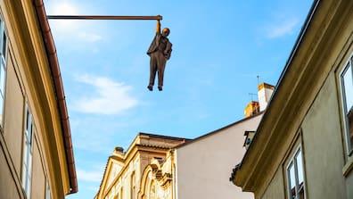 A figure represents a man hanging from a building roof with only one hand on a rod.