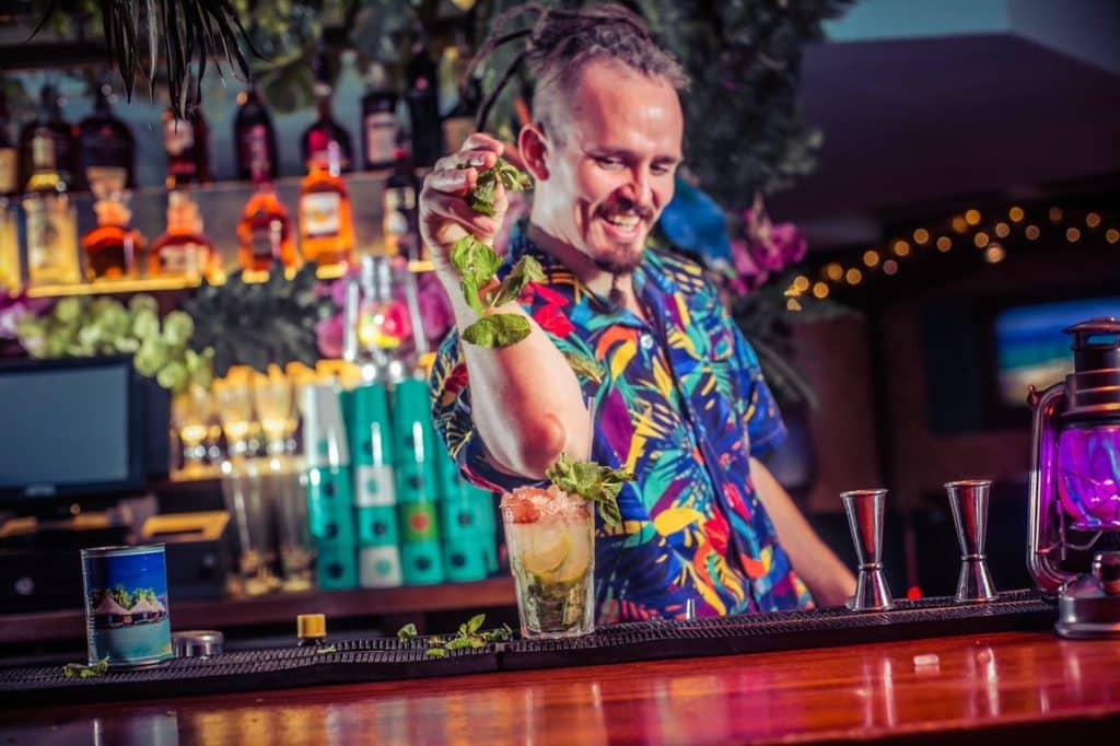 A member of the barrio bar team in a brightly coloured shirt dropping a garnish onto a cocktail at the bar